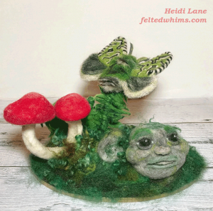 Felt sculpture of two red mushrooms, a green moth on a fern and a humanoid face on the mossy ground