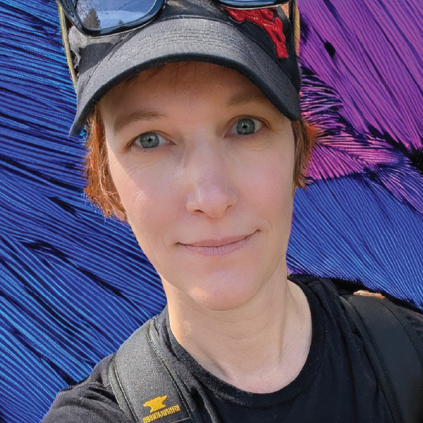 Short-haired person in black baseball hat and black T-shirt in front of blue, pink and violet background.