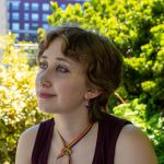 pale skinned person wearing a rainbow ribbon around their neck. Green foliage in background.